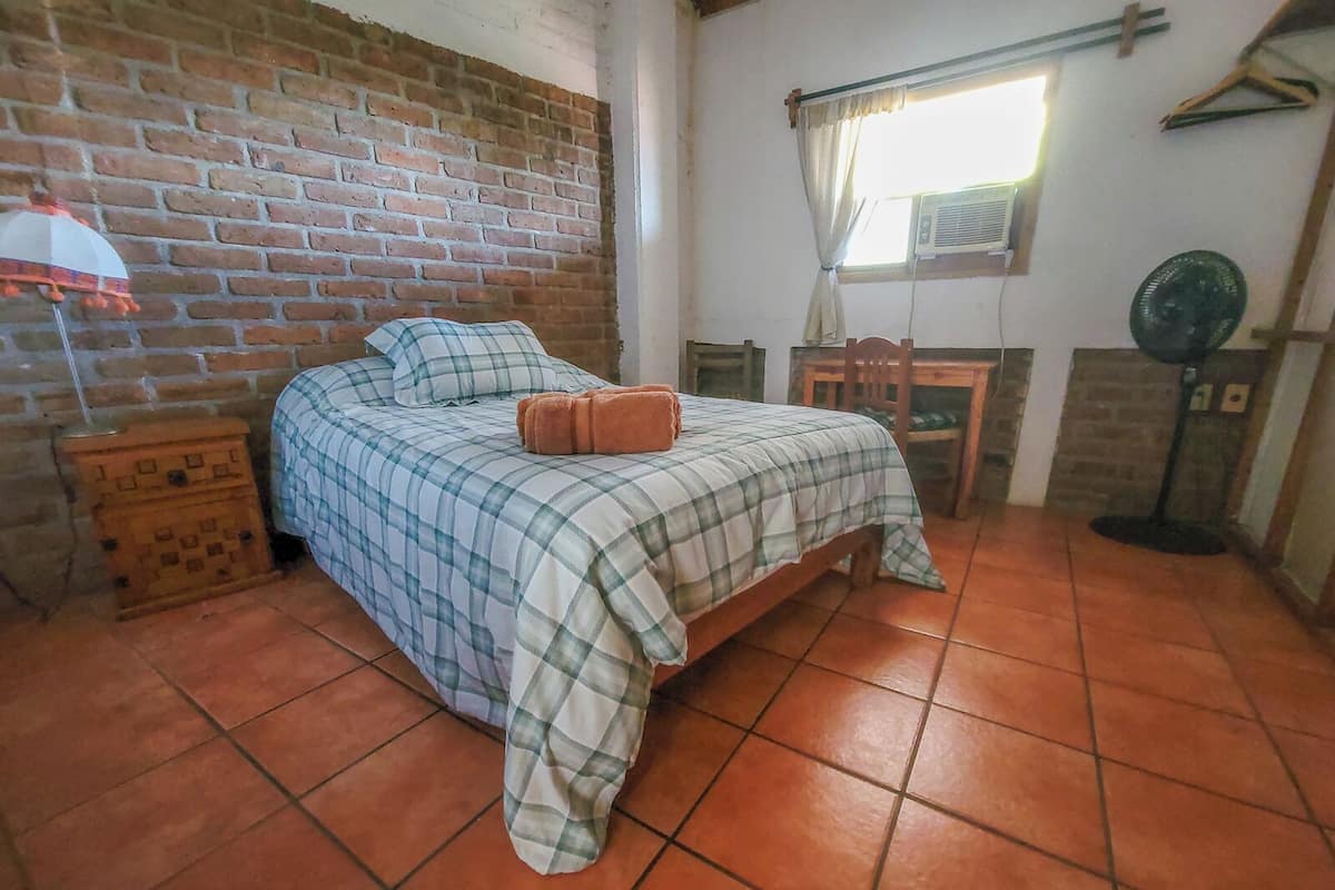 A room with a brick wall, a bed with a checkered bedspread, a wooden nightstand, a table with chairs, a window with curtains, and a standing fan.