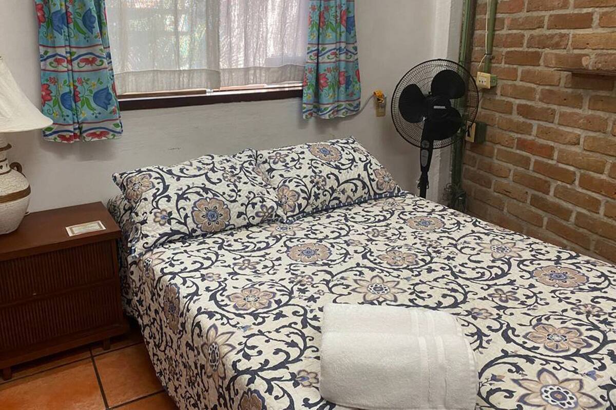 A bedroom with a patterned bedspread, a bedside lamp, a fan, and a window with curtains.