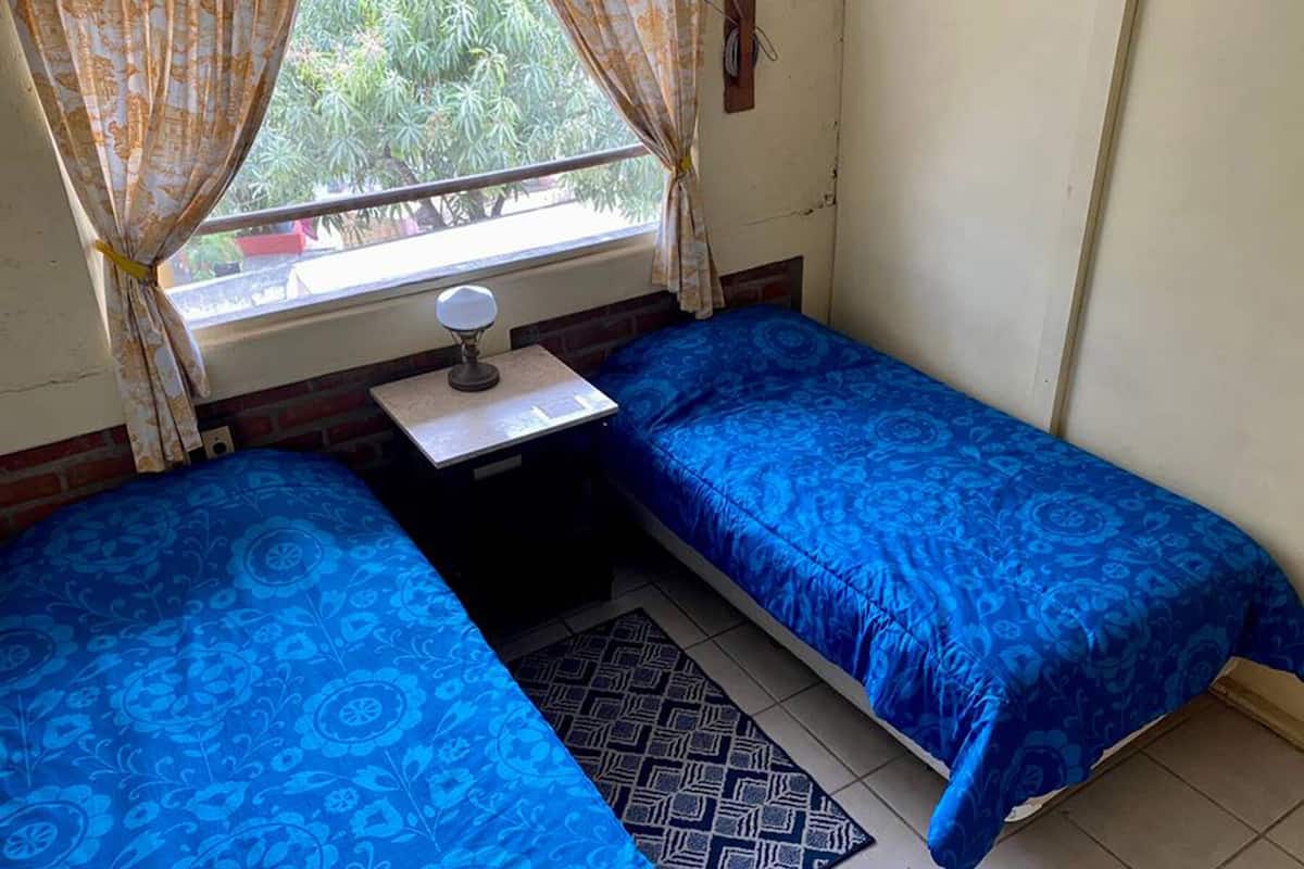 A small room with two single beds, a desk with a lamp, a window with curtains, and a patterned rug.