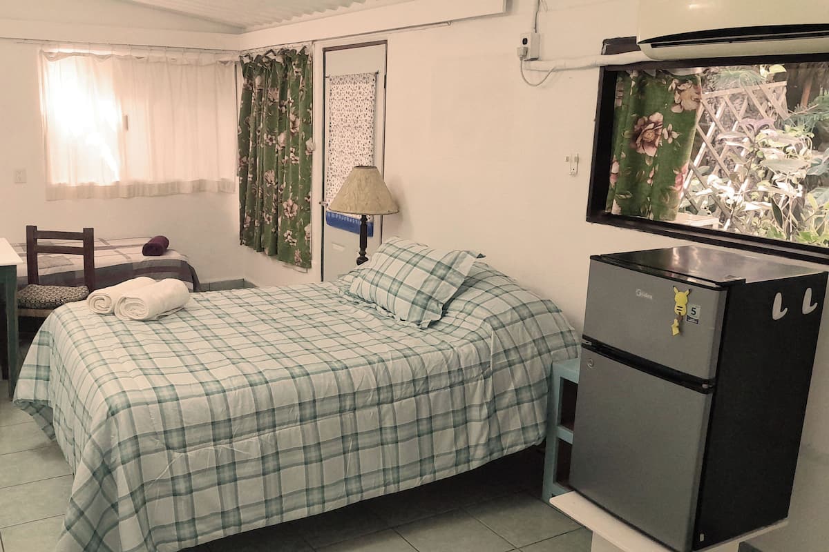 A small, tidy room with a bed, a desk, a chair, a lamp, and a refrigerator.