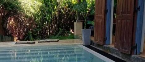 A swimming pool with a wooden deck and a covered area with blue shutters.