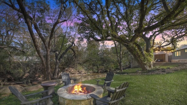 An outdoor fire pit area with seating, surrounded by trees and a house in the background. - Guest Quarters - 2 Blocks to Main Street! (Fredericksburg)