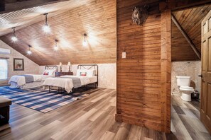 Een gezellige, rustieke slaapkamer met houten plafonds en muren, twee bedden, een groot raam, en een badkamer met toilet en wastafel.