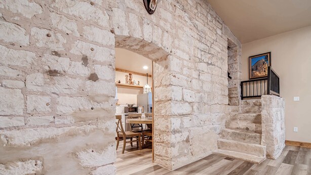 A stone-walled interior with a dining area, a staircase, and a view of the outside.