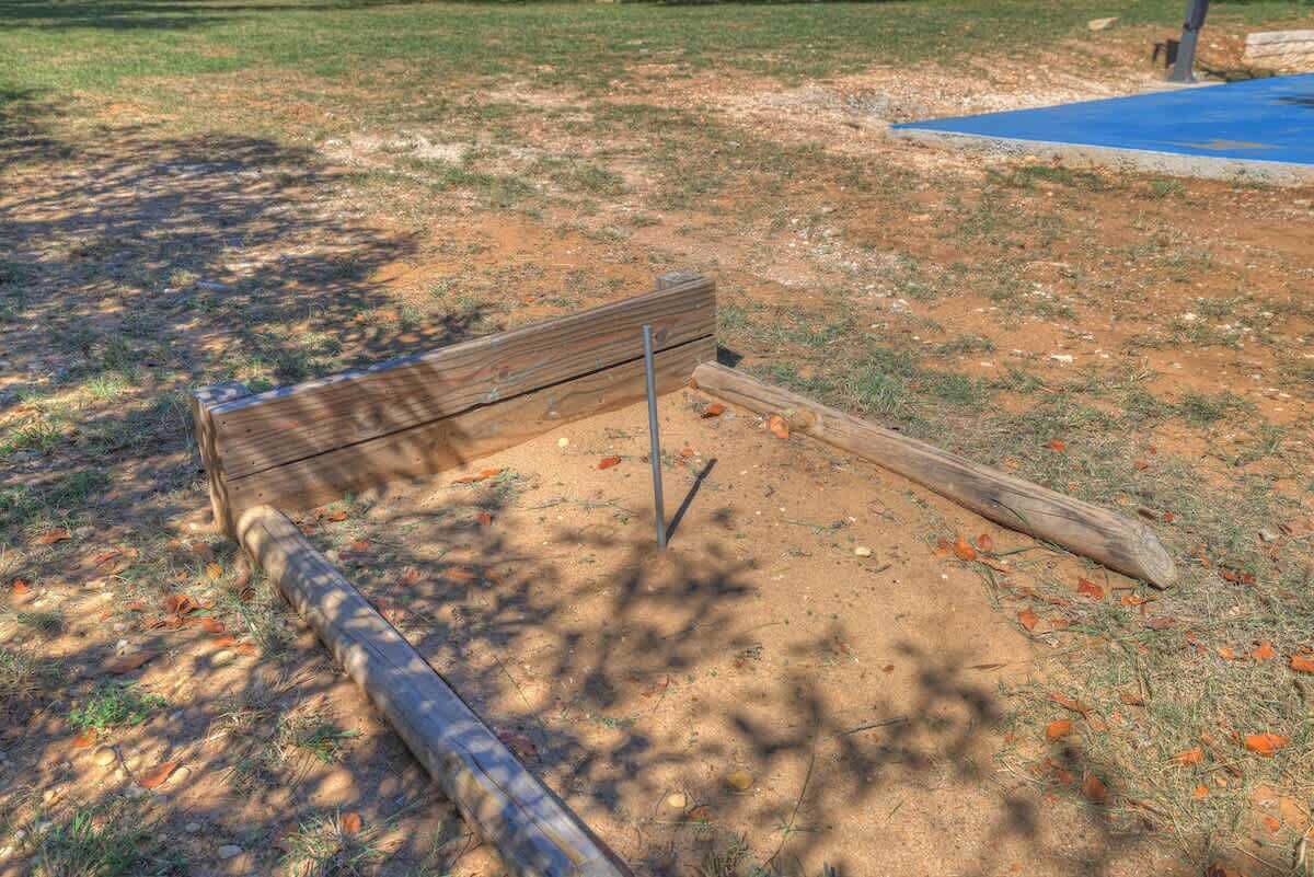 Un banco de madera en un parque con suelo de tierra y hierba escasa.