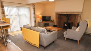 A cozy living room with a sofa, armchair, and a small table. There is a fireplace, a television, and a dining area with a table and chairs. - Henhill Lodge (Chathill)
