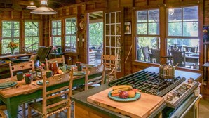 A rustic cabin interior with a dining area, wooden furniture, and large windows. - Private Lakehouse with Great Swimming and Epic Sunset Views (Colchester)
