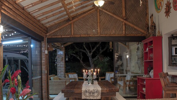 Un espacio habitable amplio con techo alto, vigas de madera, un sofá con estampados tropicales, una mesa de comedor con mantel de encaje y una estantería roja.