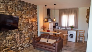 A compact living space with a stone wall, a brown leather sofa, a flat-screen TV, a kitchen with a washing machine, and a dining area. - CASAS RURALES MORADA DEL CERRO (Alcala del Jucar)