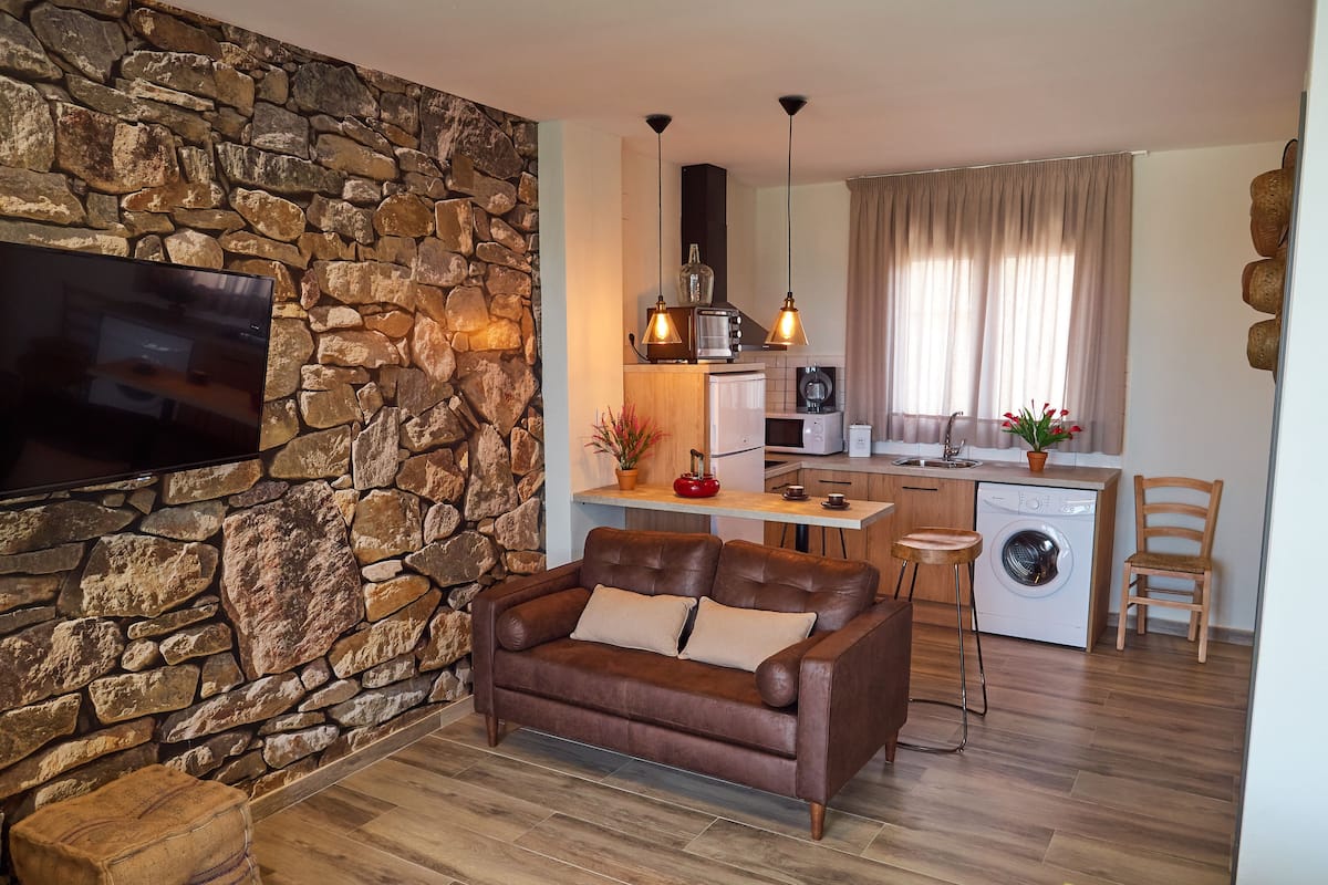 A compact living space with a stone wall, a brown leather sofa, a flat-screen TV, a kitchen with a washing machine, and a dining area.