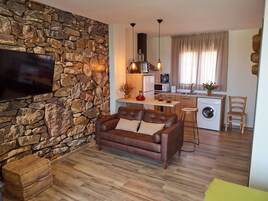 A compact living space with a stone wall, a brown leather sofa, a flat-screen TV, a kitchen with a washing machine, and a dining area.
