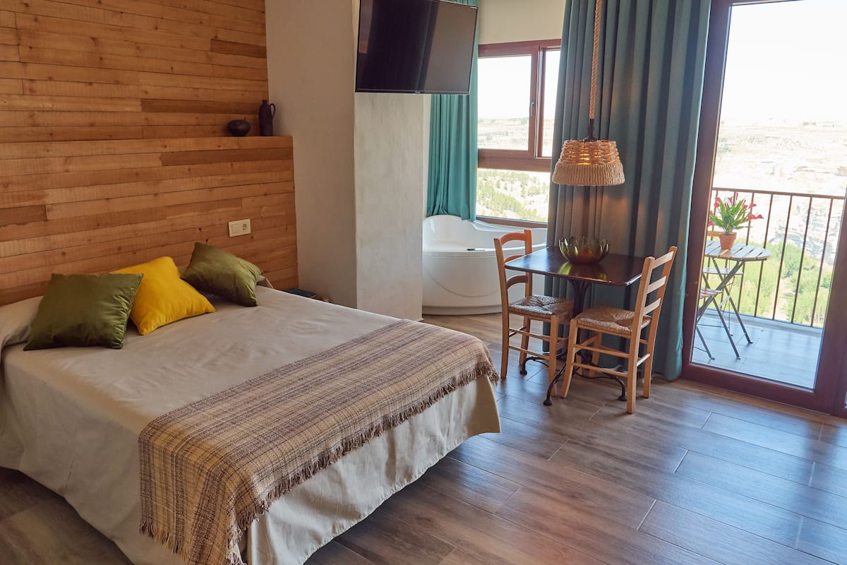 A bedroom with a bed, a table with chairs, a TV, and a balcony with a view.