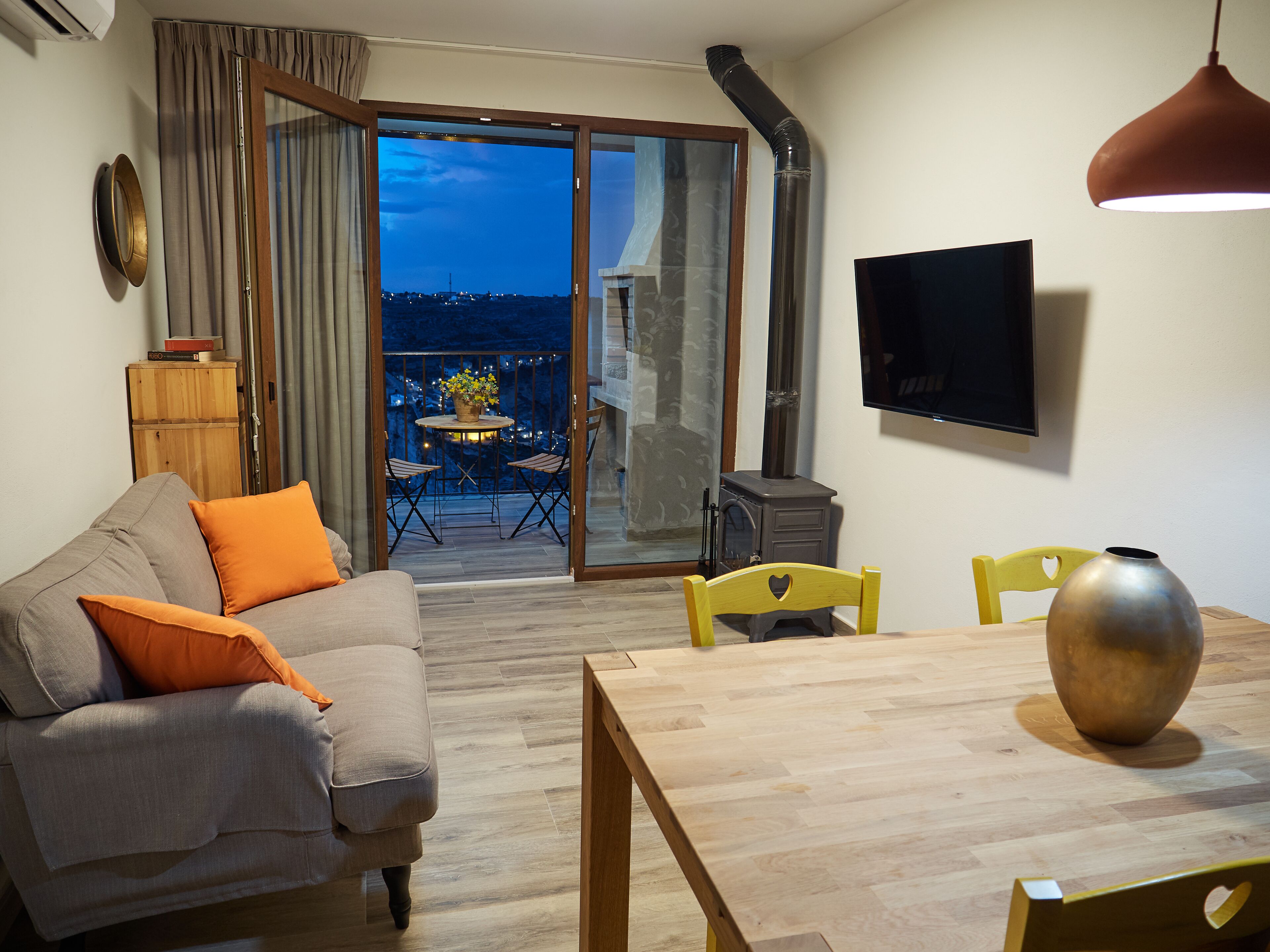 Sala de estar com sofá, mesa, cadeiras, televisão na parede e porta de vidro deslizante que dá para uma varanda externa com vista da cidade à noite.