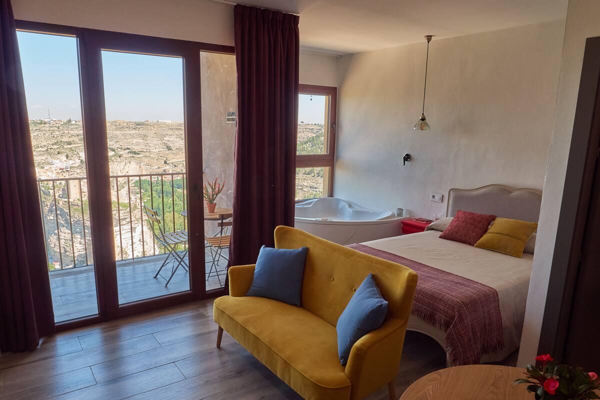 A hotel room with a balcony, a bed, a sofa, a table, and a potted plant.