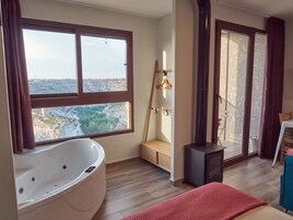 A modern bathroom with a large bathtub, a wooden bench, and a view of a cityscape through large windows.