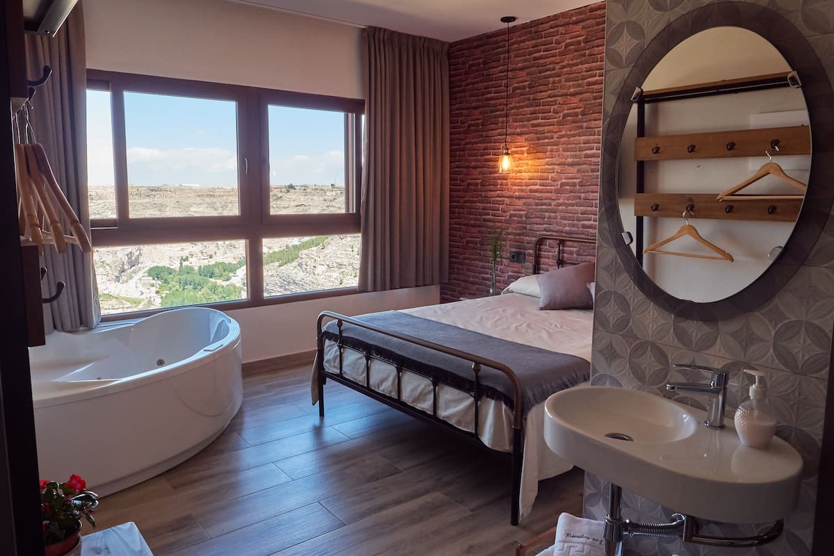 A bedroom with a large window, a bathtub, a bed, and a bathroom with a sink and mirror.