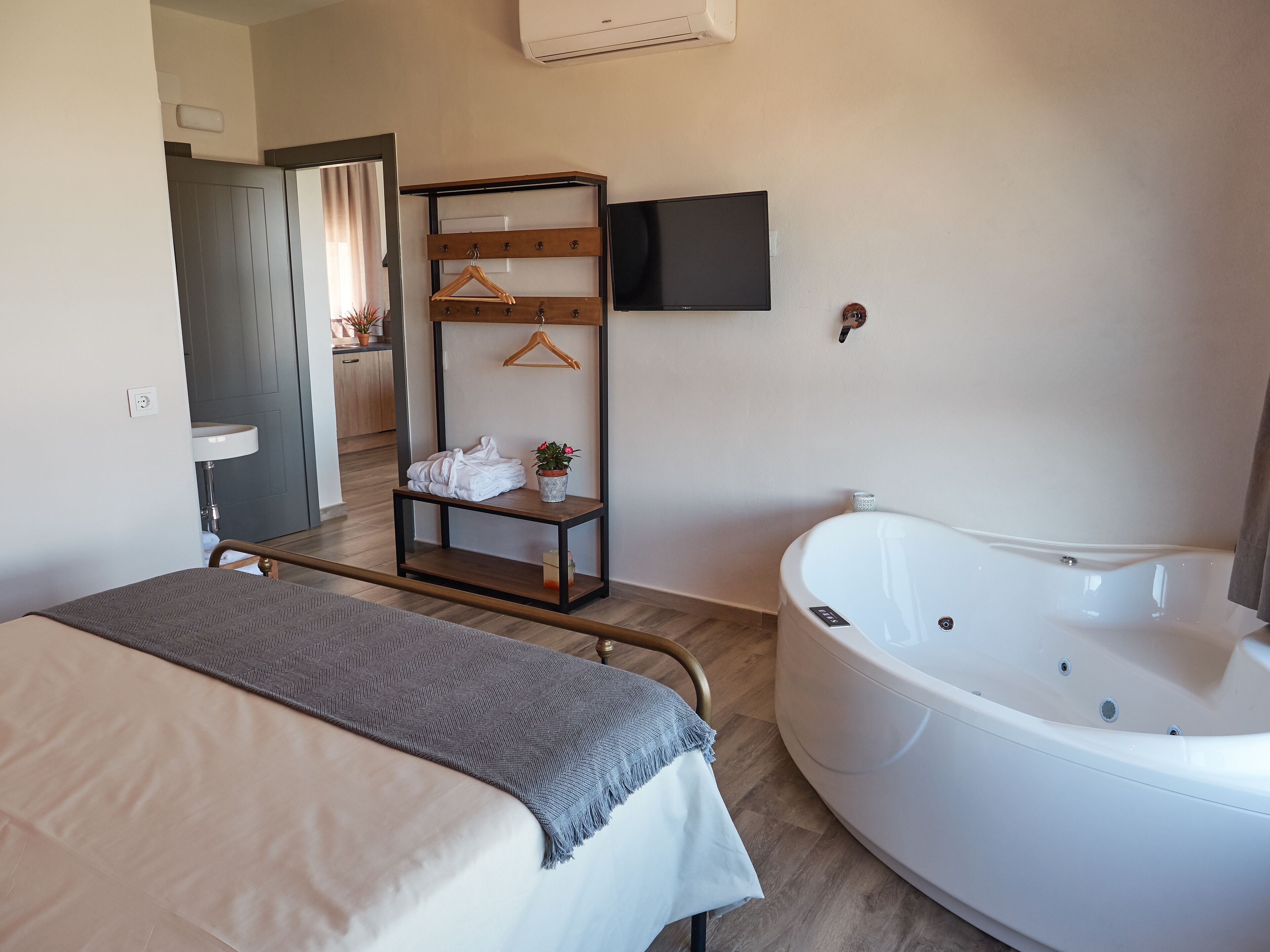 A modern bedroom with a bed, a bedside table with a towel and a potted plant, a wooden shelf with hangers, a flat-screen TV, and a large white bathtub.