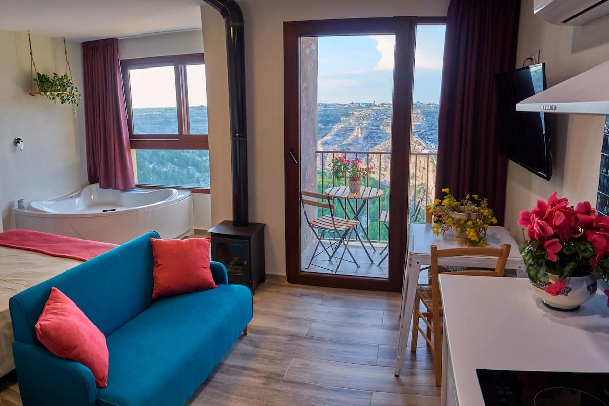 A bedroom with a large bed, a blue sofa, a bathroom with a bathtub, and a balcony with a table and chairs.