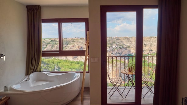 A bedroom with a large bathtub, a balcony with a table and chairs, and a view of a rocky landscape. - CASAS RURALES MORADA DEL CERRO (Alcala del Jucar)