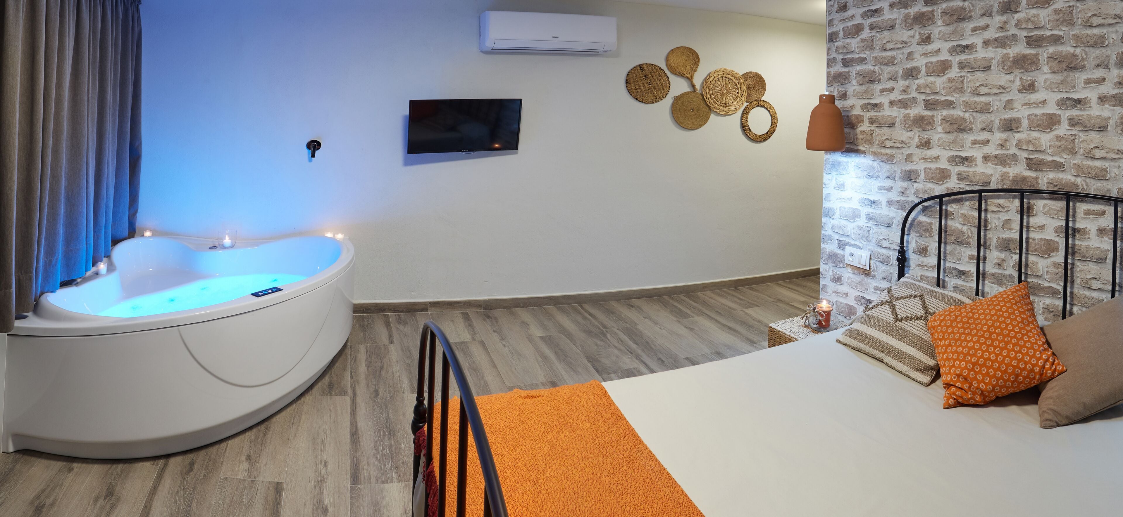 A modern room with a curved bathtub, a bed with orange bedding, a brick wall, and a mounted TV.