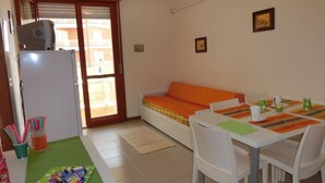 A small, well-lit room with a kitchenette, a sofa, a dining table with chairs, and a view through a sliding door. - Seaside Serenity in our Flat (San Michele al Tagliamento)