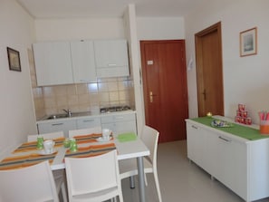 A compact kitchen with white cabinets, a dining table with chairs, and a small countertop with food items.