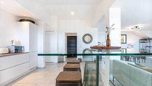 A modern kitchen with white cabinetry, a glass-top dining table, and wooden stools. - Spacious villa w/ exceptional sea-view, pool & petanque ground _ 5 mn from beach (Lumio)