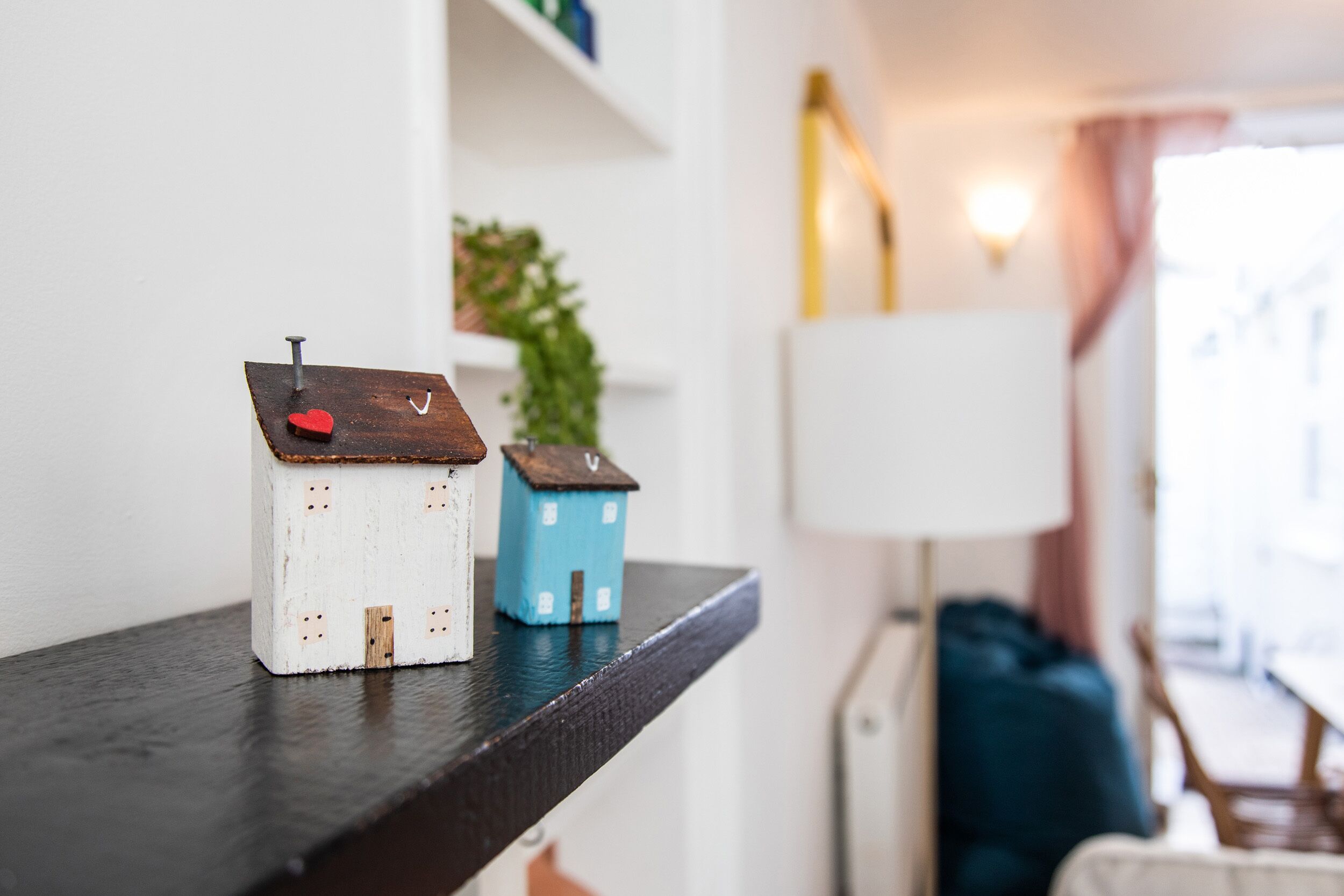 Une étagère avec deux petites maisons au style rustique, une lampe et une plante, dans une pièce dont le mur est blanc et qui possède une fenêtre.