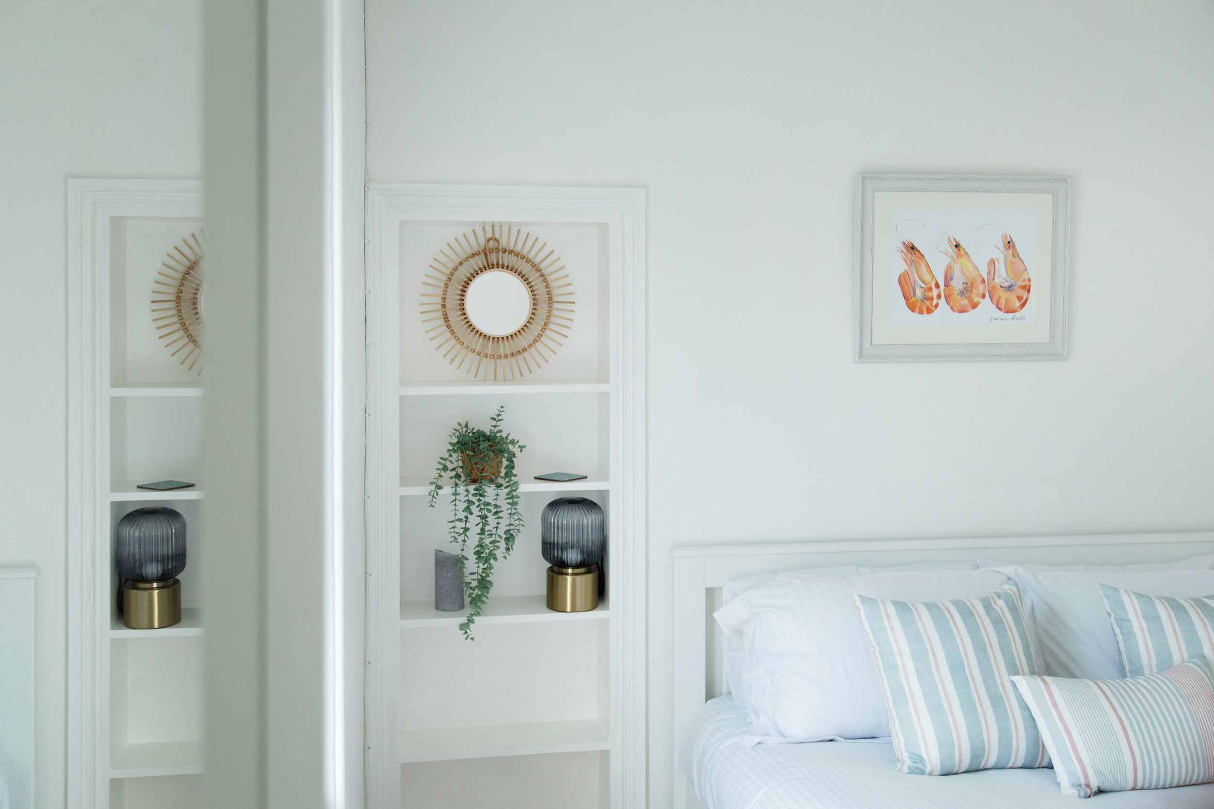 Un dormitorio con cama blanca, almohadas a rayas, una obra de arte enmarcada de camarones y dos elementos decorativos fijados a la pared.