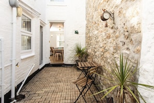 A narrow courtyard with a brick floor, a wooden table, and chairs. There is a white wall with a stone texture and a wall-mounted clock.