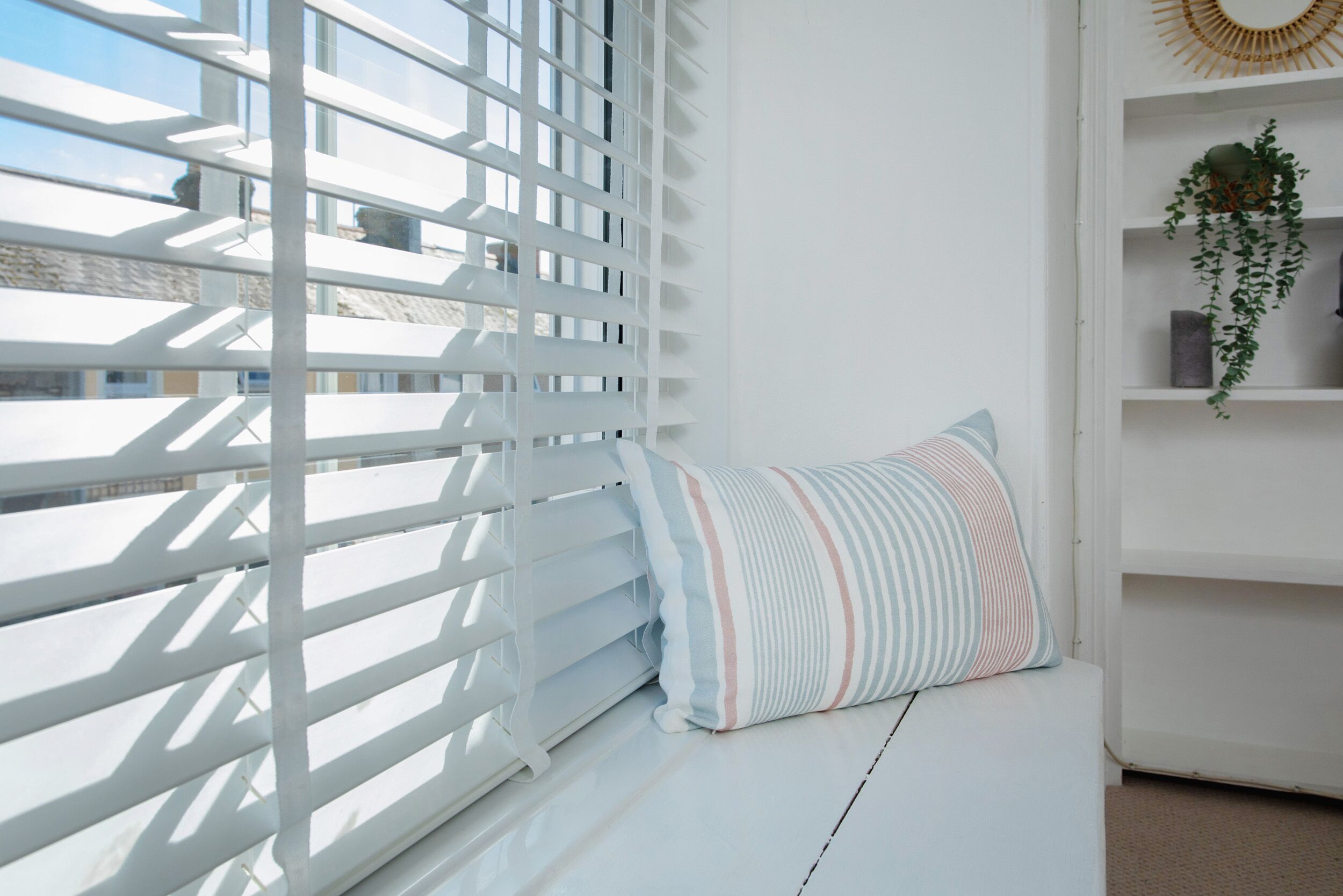 A white room with a window covered by white blinds, a striped pillow on a white bench, and a shelf with a plant and a decorative item.