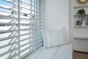 A white room with a window covered by white blinds, a striped pillow on a white bench, and a shelf with a plant and a decorative item. - 11 Belgravia Street (Penzance)