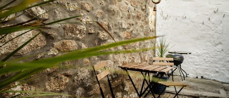 A rustic outdoor seating area with a stone wall, wooden table and chairs, a clock, and a barbecue grill.