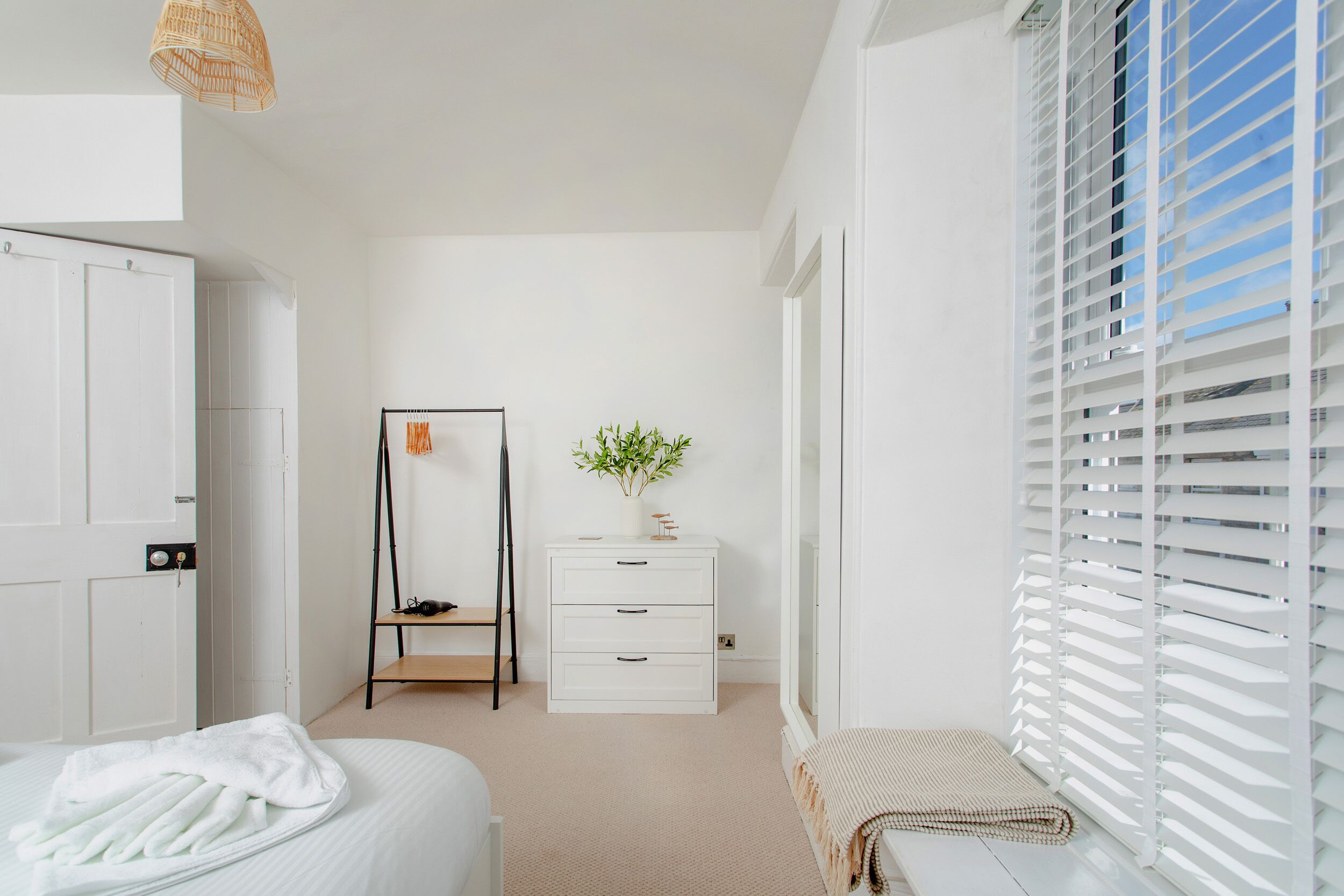 A minimalist bedroom with a bed, a dresser, a shelf with shoes, a plant, and a window with blinds.