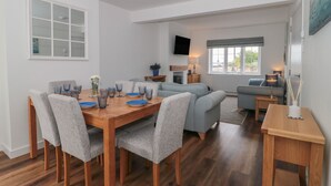 A modern living room with a wooden dining table, grey upholstered chairs, a grey sofa, and a television mounted on the wall. - Quay Cottage - Sea View (Poole)