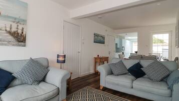 A spacious living room with two grey sofas, patterned cushions, a wooden coffee table, and a patterned rug. A painting of a beach scene is on the wall, and a door leads to a kitchen area.