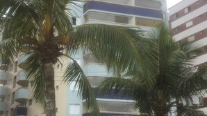 A multi-story residential building with balconies, a gated entrance, and palm trees in the foreground. - Foot in the sand at Caiçara beach II (Praia Grande)