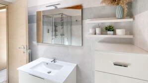 Ein modernes Badezimmer mit einer in Glas abgeschirmten Dusche, einem weißen Waschbecken und einem Spiegel darüber. Über dem Waschbecken befinden sich Ablageflächen und auf der linken Seite eine Holztür.