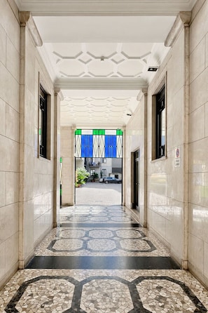 Een gang met een patroon in de vloer, een plafond met geometrisch ontwerp en een uitzicht op een gebouw met een blauw-witte gestreepte luifel.
