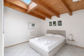 Une chambre avec un grand lit, des poutres apparentes en bois au plafond, des murs blancs et une petite table de chevet avec une lampe.