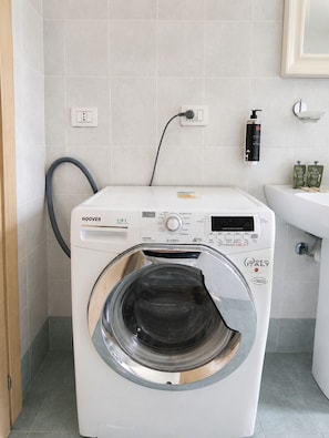 A front-loading white washing machine with a control panel, a power outlet, and a wall-mounted soap dispenser. - BIG Suite Goldoni [Duomo-M4 Lin Airport] (Milan)