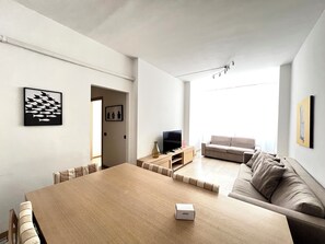 A modern living room with a wooden dining table, a sofa, a TV, and artwork on the walls.