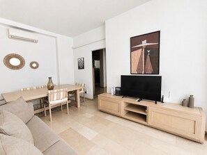 A modern living room with a flat-screen TV, a dining area with a wooden table and chairs, and a beige sofa.