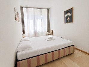 A single bed with a white mattress and striped bedding, a chair, a bedside table with a lamp, and a framed picture on the wall.