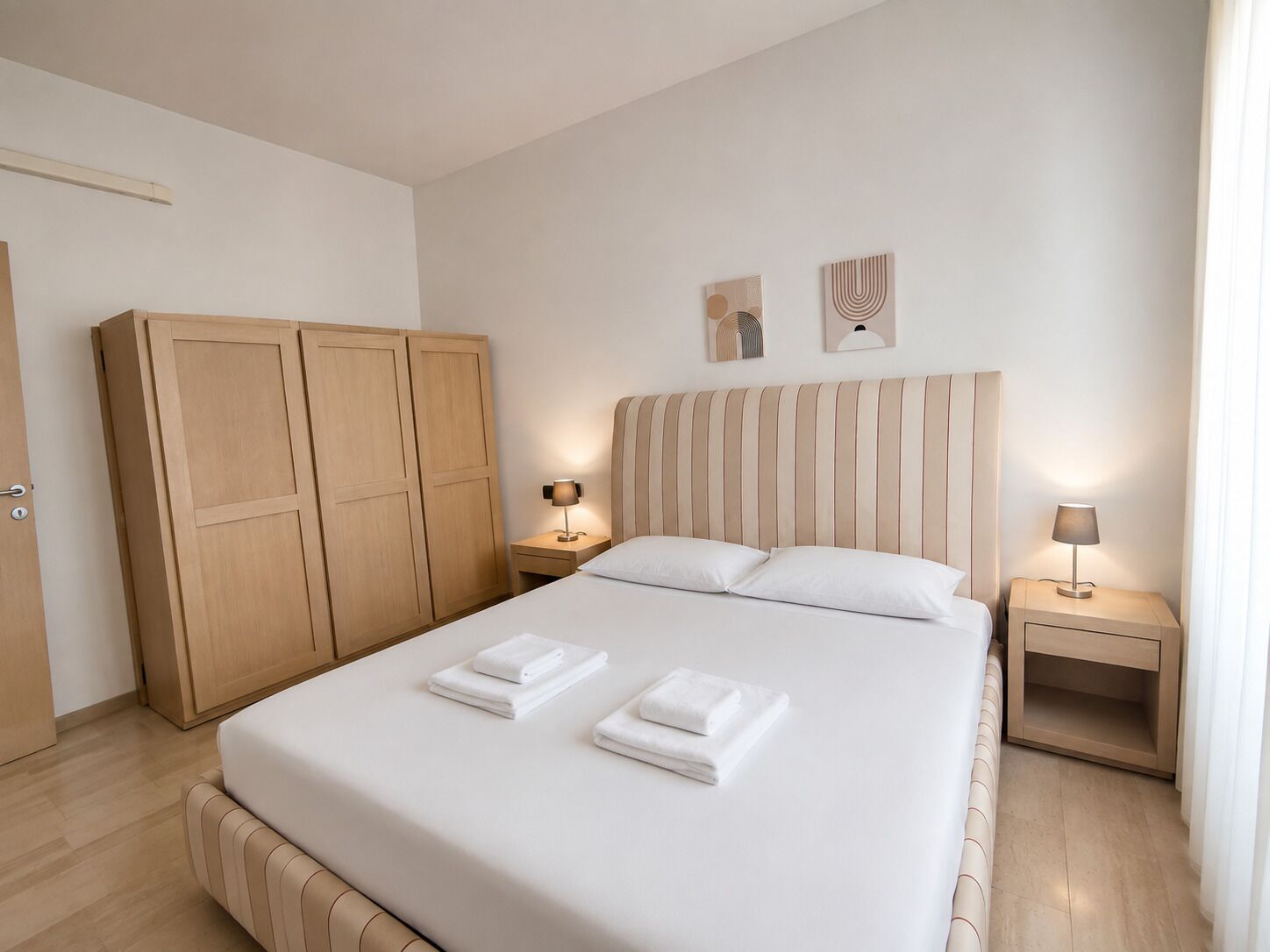 A hotel room with a large bed, two bedside tables, a wardrobe, and two wall art pieces.