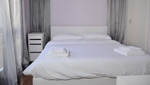 Una cama bien tendida con sábanas y toallas blancas, una mesita de noche blanca y cortinas grises.