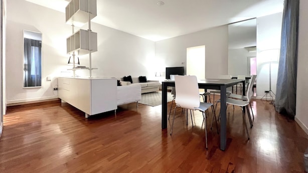 Una sala de estar moderna y bien iluminada con un sofá blanco, una mesa de comedor con sillas y un espejo grande.