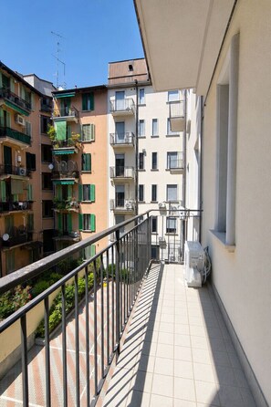 Un balcón estrecho con barandilla metálica, que da a una hilera de edificios de varios pisos con balcones y ventanas.