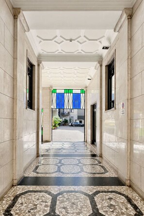 A corridor with a patterned floor, a ceiling with a geometric design, and a view into a building with a blue and white striped awning.