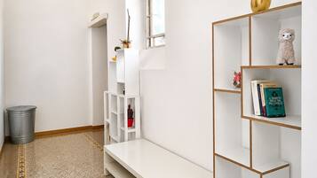 Un couloir minimaliste avec un banc blanc, une poubelle grise et une étagère blanche contenant des livres et des objets décoratifs.